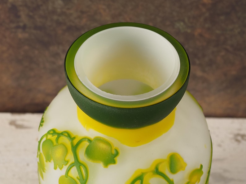 Puede incluir: Jarr&oacute;n de vidrio blanco esmerilado con un borde verde y amarillo. El jarr&oacute;n est&aacute; decorado con un patr&oacute;n floral de flores de lirio de los valles verdes.