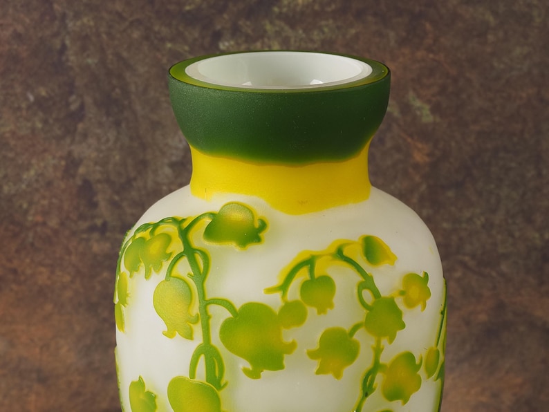 Puede incluir: Un jarr&oacute;n de vidrio blanco con un borde verde y amarillo. El jarr&oacute;n est&aacute; decorado con un patr&oacute;n floral de lirios del valle.