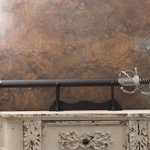 Puede incluir: Una espada decorativa con una vaina negra y una empuñadura plateada ornamentada, apoyada sobre un soporte negro. La empuñadura presenta diseños intrincados y un mango envuelto. La espada se exhibe sobre un mueble blanco con detalles tallados sobre un fondo marrón texturizado.