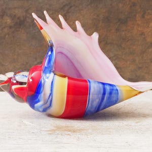 Può includere: Scultura di conchiglia in vetro colorato. La conchiglia presenta un motivo a strisce rosa, rosse, blu e gialle. L'oggetto è posto su una superficie bianca.