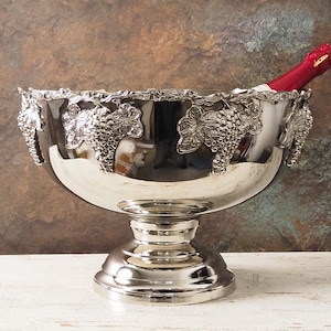 May include: A silver wine chiller with a pedestal base, adorned with grape vine detailing around the rim. A red wine bottle is partially visible inside. The chiller sits on a white surface against a textured brown and blue background.