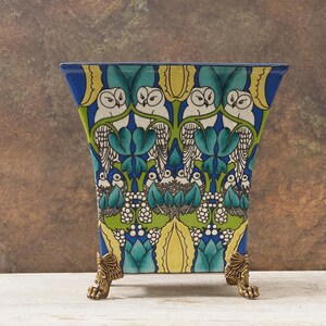Peut inclure: Un vase décoratif en céramique avec un bord bleu et des pieds en laiton. Le vase présente un motif coloré de hiboux, de feuilles et de motifs floraux dans des tons de bleu, vert, jaune et blanc. Un objet de décoration intérieure unique.
