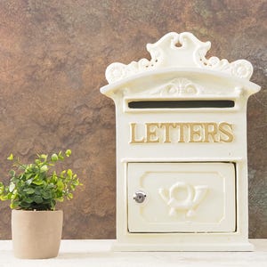 May include: Cream-colored mailbox with the word "LETTERS" in gold lettering. The mailbox has a decorative top and a small door with a keyhole. A small potted plant with green leaves sits to the left of the mailbox.