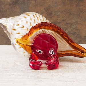 Puede incluir: Una escultura de arte en vidrio de un cangrejo rojo que emerge de una concha. El cangrejo tiene pinzas y ojos detallados. La concha es blanca con detalles dorados y un borde marrón y naranja.