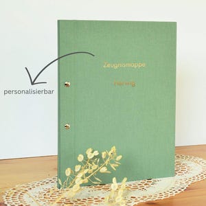 Op de afbeelding: Een saliegroene map met gouden letters met de tekst "Zeugnismappe norwig". De map heeft drie gouden klinknagels en is personaliseerbaar. Een witte theekop met een zilveren rand staat op een kanten kleedje.