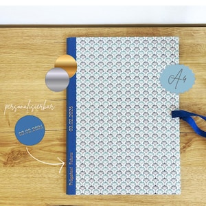 Puede incluir: Un diario estampado con lomo y cinta azules, con un diseño repetitivo en forma de abanico en verde azulado y gris sobre fondo blanco. El diario tiene una etiqueta azul claro con las letras "Aa" y el texto "03.02.2026" en el lomo.
