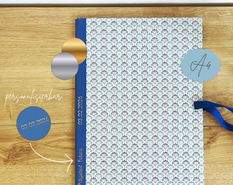 May include: A patterned journal with a blue spine and ribbon, featuring a repeating fan-like design in teal and gray on a white background. The journal has a light blue tag with the letters "Aa" and the text "03.02.2026" on the spine.