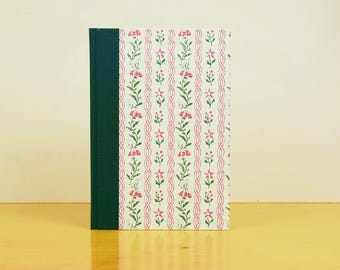 May include: A hardcover journal with a green spine and a floral pattern. The cover features a white background with pink flowers and green leaves arranged in vertical rows. The journal is standing upright on a wooden surface.