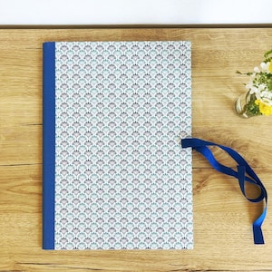 Könnte beinhalten: Ein Notizbuch mit blauem Rücken und passendem blauem Band. Das Cover zeigt ein sich wiederholendes Fächerdesign in Türkis, Blau und Grau auf Weiß. Eine kleine Vase mit gelben Blumen steht daneben. Das Notizbuch liegt auf einer Holzoberfläche.