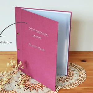 Può includere: Un raccoglitore di documenti magenta con testo argentato "DOKUMENTEN MAPPE" e "Familie Bauer". Il raccoglitore è aperto, rivelando pagine bianche. Sono visibili anche le parole "personalizzabile".