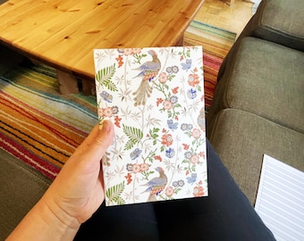 May include: A notebook with a white background featuring a colorful floral and bird pattern. The design includes blue, orange, and green elements. The notebook is held in a person's hand.