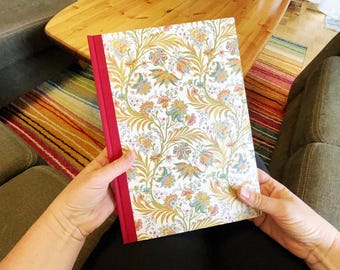 May include: A floral patterned notebook with a red spine. The cover features a white background with a repeating design of gold, blue, and orange flowers and leaves. The notebook is held open by a person.