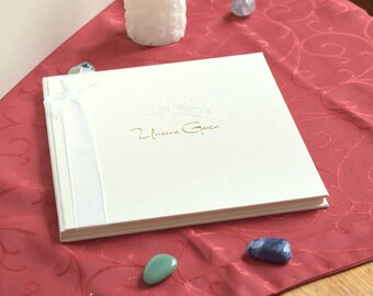 May include: A rectangular white guest book with a smooth cover featuring the gold embossed text "Unsere Gäste" and a small bird illustration. A thin white ribbon is wrapped vertically around the left side of the book, which is closed and lying flat.