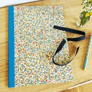 Op de afbeelding: Een decoratief notitieboek met een bloemmotief in blauw, groen en goud, gebonden met een helderblauwe rug. Een zwart lint is om het notitieboek gebonden. Een lichtblauwe potlood en een kleine vaas met bloemen liggen ook op het houten oppervlak.