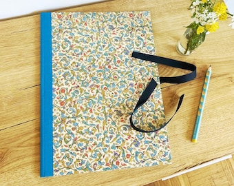May include: A decorative journal with a floral pattern in shades of blue, green, and gold, bound with a bright blue spine. A black ribbon is tied around the journal. A light blue pencil and a small vase of flowers are also on the wooden surface.
