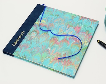 May include: A hardbound guest book with a blue cover and a patterned design in shades of blue, pink, and green. The spine has the word "Gästebuch" in gold lettering. A blue ribbon is attached. A black pen and a teal ceramic cup are also visible.