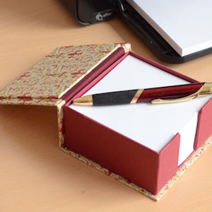 May include: A red and gold pen lies on a white notepad within a decorative box. The box is red with a patterned lid and base. The pen has a black grip and gold accents. The notepad is square.