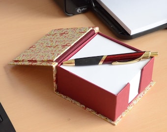 May include: A red and gold pen rests on a white notepad inside a decorative box. The box is red with a patterned lid and base. The pen has a black grip and gold accents. The notepad is square.