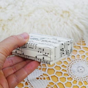 Könnte beinhalten: Eine rechteckige Schachtel mit Notenblättern. Die Schachtel ist weiß mit schwarzen Noten und Notenlinien. Die Schachtel wird in einer Hand gehalten, mit einem weichen, weißen, flauschigen Hintergrund.