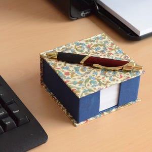 Può includere: Un portablocco per appunti quadrato con base blu e coperchio caratterizzato da un motivo floreale color crema, blu e rosso. Una penna a sfera retrattile con fusto rosso scuro, impugnatura nera e dettagli dorati poggia sul coperchio chiuso.