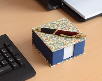 May include: A square memo pad holder with a blue base and a lid featuring a cream, blue, and red floral pattern. A retractable ballpoint pen with a dark red barrel, black grip, and gold-tone accents rests horizontally on top of the closed lid.