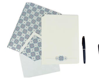 May include: Stationery set featuring a cream-colored notepad with a decorative blue and white floral design, matching envelope, and patterned paper. A black pen is placed to the right of the notepad.