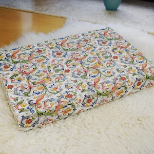 Peut inclure: Une boîte rectangulaire ornée d'un motif floral coloré rouge, jaune, bleu et vert sur fond blanc. La boîte repose sur un tapis blanc et duveteux.