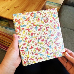 Könnte beinhalten: Ein quadratisches Notizbuch mit cremefarbenem Einband und einem bunten Blumenmuster in Rot, Gelb, Blau und Grün. Das Notizbuch wird vor einem Holztisch und einem grünen Sofa gehalten.