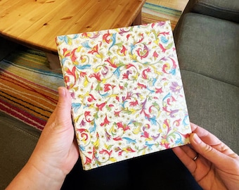 May include: A square notebook with a cream-colored cover featuring a colorful floral pattern in red, yellow, blue, and green. The notebook is held in front of a wooden table and a green couch.