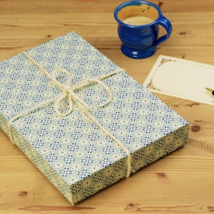 Pode incluir: Uma caixa retangular embrulhada em papel com padrão azul, branco e dourado, atada com cordel branco, acompanhada por uma caneca de cerâmica azul cheia de café, um cartão retangular em branco com borda decorativa e uma caneta-tinteiro preta.