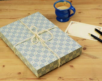May include: A rectangular box wrapped in blue, white, and gold patterned paper tied with white twine, accompanied by a blue ceramic mug filled with coffee, a blank rectangular card with a decorative border, and a black fountain pen.