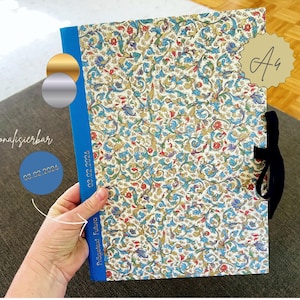 Puede incluir: Un cuaderno con una cubierta estampada floral, lomo azul y cierre de cinta negra. La cubierta presenta un diseño colorido con acentos azules, rojos y dorados. El texto "02.02.2026" está impreso en el lomo. El cuaderno está etiquetado "A4".