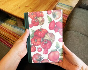 May include: A notebook with a green spine and a cover featuring a watercolor illustration of various tomatoes and pumpkins. The design includes red, orange, and green hues, with detailed depictions of the vegetables.