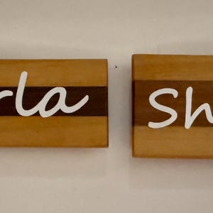 May include: Two wooden nameplates, one reading "Marla" and the other "Shari", are displayed. Each name is in white cursive script, set against a light brown wood background with a dark brown stripe.