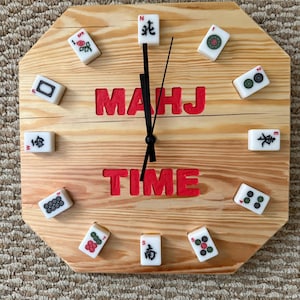 May include: Wooden octagonal clock with the words "MAHJ TIME" in red. The clock face is made of light-colored wood and features Mahjong tiles as hour markers. Black clock hands indicate the time.