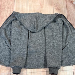 Könnte beinhalten: Ein grauer Kapuzenpullover mit durchgehendem Reißverschluss, offen, um das Innere zu zeigen. Die Bündchen und das Kapuzenfutter sind in einem dunkleren Grauton gehalten. Der Pullover ist aus weichem, strukturiertem Material.