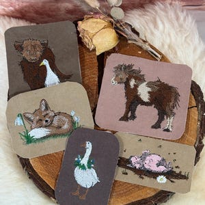Könnte beinhalten: Eine Sammlung gestickter Aufnäher mit verschiedenen Tieren. Designs umfassen ein Highland-Rind mit einer Gans, ein Pony, einen schlafenden Fuchs, eine Gans mit Blumenkranz und ein Ferkel. Die Aufnäher sind in Braun-, Beige- und Rosatönen gehalten.