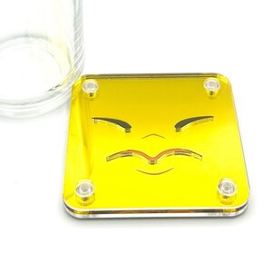 May include: A yellow square coaster with a black and orange frowning face design. The coaster is made of acrylic and has a glossy finish.