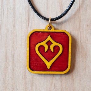 May include: A red wooden pendant with a gold heart design hangs from a black cord necklace. The pendant is square shaped with rounded corners.