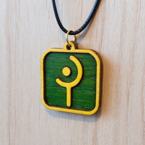 May include: A green wooden pendant with a gold outline and a gold symbol in the center. The pendant is hanging from a black cord.