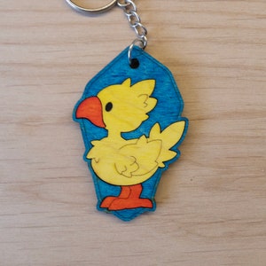 May include: A yellow and orange cartoon bird keychain with a blue background. The keychain is attached to a silver key ring.