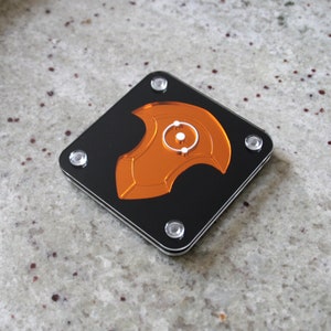 Puede incluir: Un posavasos cuadrado de acr&iacute;lico negro con un dise&ntilde;o naranja estilizado en el centro. El dise&ntilde;o presenta un elemento circular con un c&iacute;rculo blanco m&aacute;s peque&ntilde;o en el interior. Cuatro tornillos plateados aseguran el posavasos.