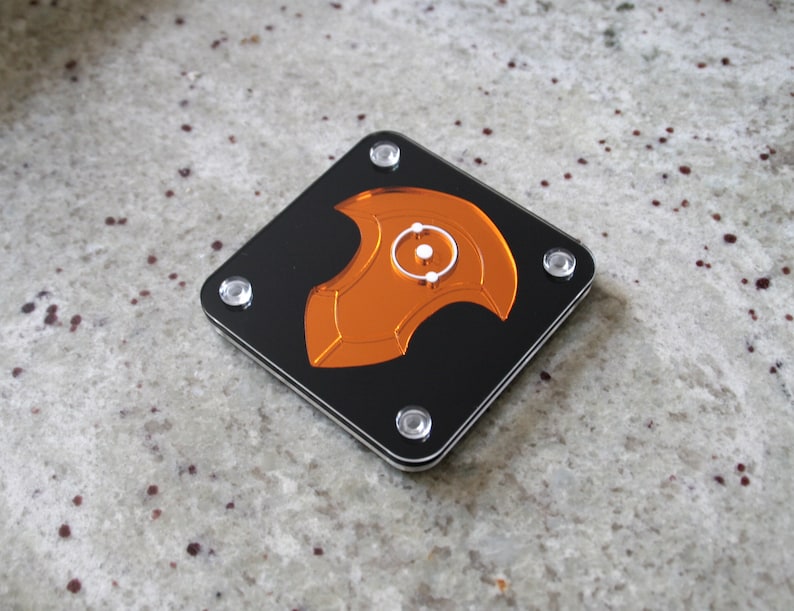 Puede incluir: Un posavasos cuadrado de acr&iacute;lico negro con un dise&ntilde;o naranja estilizado en el centro. El dise&ntilde;o presenta un elemento circular con un c&iacute;rculo blanco m&aacute;s peque&ntilde;o en el interior. Cuatro tornillos plateados aseguran el posavasos.