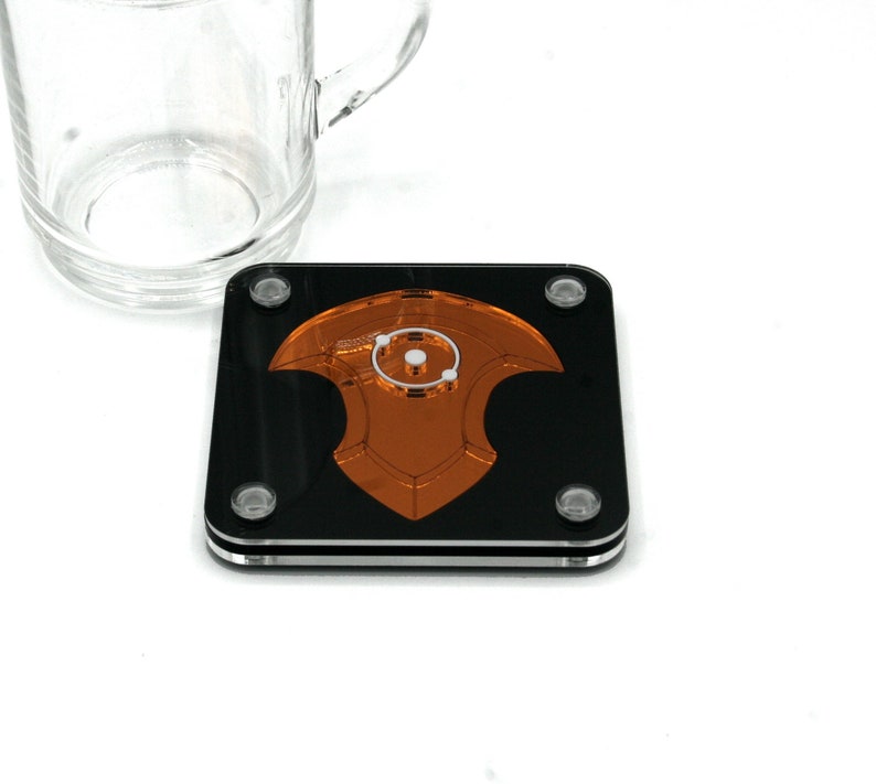 Puede incluir: Posavasos cuadrado negro y naranja con un dise&ntilde;o decorativo. El posavasos tiene un borde transparente y peque&ntilde;os pies transparentes. Una taza de vidrio transparente est&aacute; en el fondo.