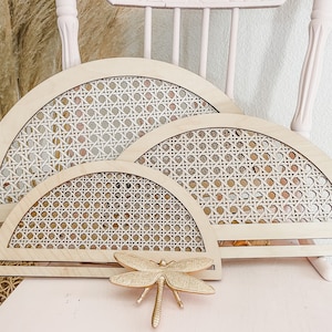 Könnte beinhalten: Drei halbkreisförmige Wanddekorationen aus Holz mit einem geflochtenen Rattanmuster. Die Stücke sind übereinander gestapelt, wobei das größte Stück unten liegt. Vor den Stücken befindet sich eine goldene Libelle.