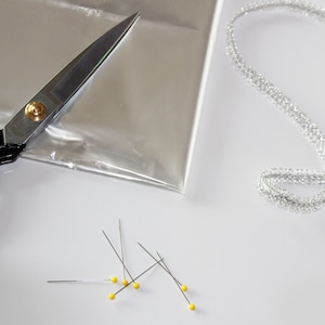 May include: A collection of craft supplies on a white surface. Includes a pair of black-handled scissors, a clear plastic sheet, a silver ribbon, and several pins with yellow heads. The items are arranged for crafting or sewing.