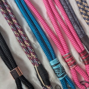 Könnte beinhalten: Nahaufnahme von mehreren farbenfrohen Schlüsselbändern. Die Schlüsselbänder bestehen aus dickem, geflochtenem Seil in verschiedenen Farben, darunter Schwarz, Pink, Blau und gemusterte Kombinationen. Einige haben Metallverschlüsse und dekorative Akzente.