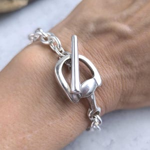 Könnte beinhalten: Ein silberfarbenes Armband mit einem Pferdezahn-Verschluss. Das Armband hat ein Kettenglied-Design und wird am Handgelenk getragen. Der Verschluss ist eine detaillierte Nachbildung eines Pferdezahns und verleiht einen einzigartigen Reitstil.