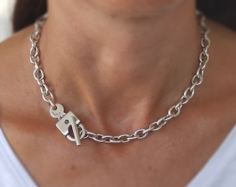 May include: A silver chain necklace with an oval link design. The necklace has a distinctive toggle clasp closure. The clasp is a square shape with a bar that slides through a loop to secure the necklace. The necklace is worn around the neck.
