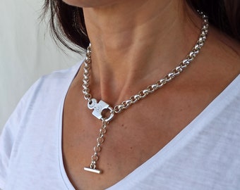 May include: A silver chain necklace with a large, square pendant that has a toggle clasp. The pendant has a detailed design and is attached to a smaller chain with a bar at the end.